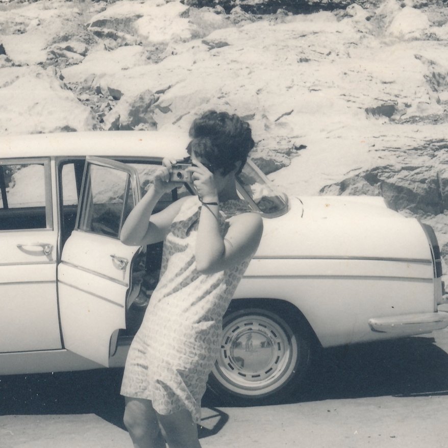 A vintage black and white photo of a woman in a patterned dress taking a picture with a camera, standing next to a classic car parked near rocky terrain.
